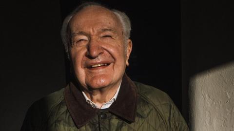 Brigadier Norman Butler, an older man with grey hair has a bright smile and wears a green coat.