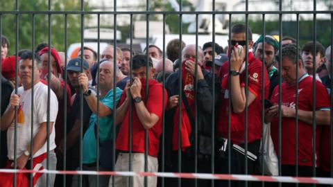 Liverpool fans queue outside the Stade de France in Paris ahead of the 2022 Champions League final. They are standing behind high metal fences.