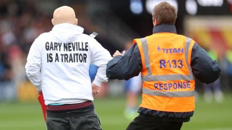 A steward chases after a man wearing a white hoodie bearing the message "Gary Neville is a traitor"