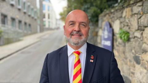 Deputy Jonathan Le Tocq - A white man with a bald head and a grey beard. He's wearing a blue suit, a white shirt, with a red and yellow tie. On the lapel of his suit is a Guernsey pin badge and a poppy. 