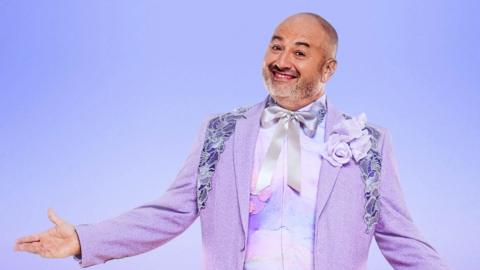 Wynne Evans in a purple suit with embroidered shoulders and a purple adn blue marble-effect waistcoat. He is grinning at the camera with one hand out. 