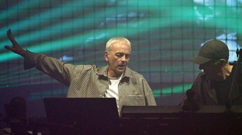Karl Hyde of Underworld performing. He has short, blond hair and is wearing a checked jacket. There are green lights illuminating the back of the stage