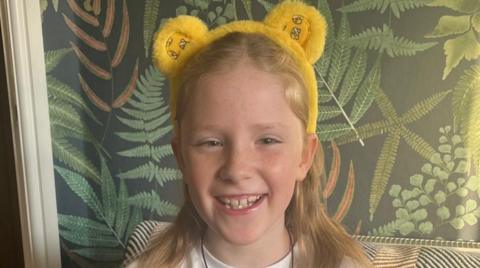 Hannah, smiling with long hair, wearing Children in Need mascot Pudsey Bear ears. She is sitting down, with a green-coloured wall behind her.