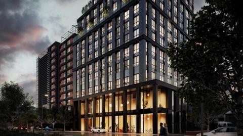 A computer generated image of a modern block of flats. The building on the image is grey with large windows at the bottom and lights inside. There are trees and greenery on the pavements where people walk and cars are driving past on the road. 