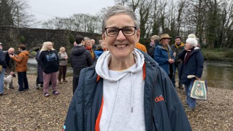 Sarah Preece, Cornwall councillor for Lostwithiel and Lanreath, on the banks of the River Fowey smiling at the camera. There are people gathered behind her.