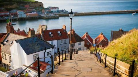 A view of Whitby from the top of the town's famous steps. The harbour is calm and has two boats in it, one heading in, the other heading out. A couple are sitting on deckchairs in the sunshine.