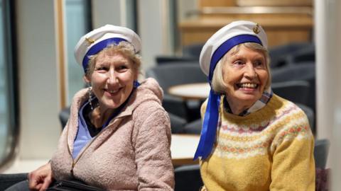 Two women on board the maiden journey of the MV Glen Sannox. They are wearing sailor hats and warm jumpers, and smiling to someone to the right of the camera. 