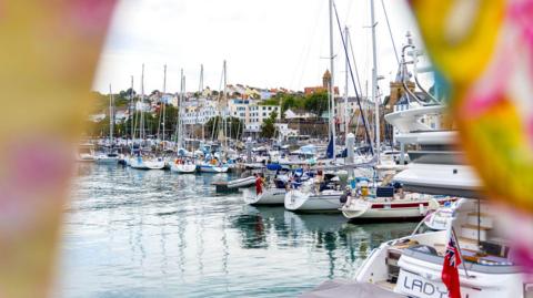 Several boats sit on the water in a marina in front of colorful houses in the background.