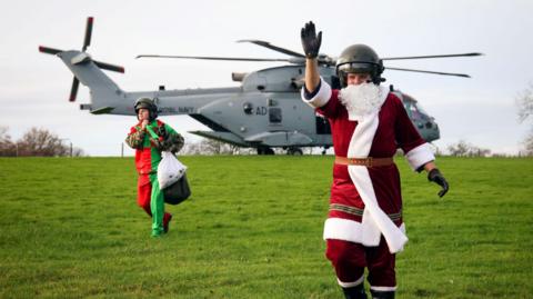 Father Christmas waving on a field with a helicopter in the background and an elf to his write.