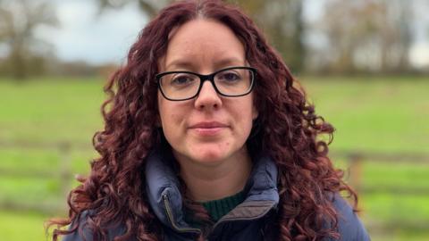 A head and shoulders shot of Kayleigh Griffiths standing in her garden looking into camera with a neutral expression. She has long reddish-brown curlyy hair and is wearing a thick navy jacket. She has black framed glasses.