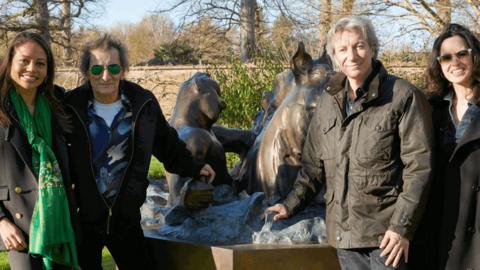 Image of two women and two men, standing in front of a bronze sculpture in a walled garden. One of the men is Ronnie Wood, who is wearing a dark jacket, sunglasses and has dark hair styled into a spiky pattern