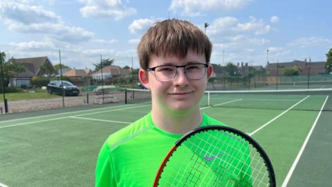 Oliver Appleby, 18, has brown hair with a fringe over his eyes. He has dark rimmed glasses and is wearing a lime coloured T-shirt. He is holding a tennis racquet. Behind him is the hard court.