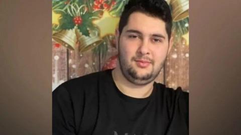 A family photo of Tsvetomir Genov, who is wearing a black t-shirt and has dark hair and facial hair. He is standing in front of a sign with Christmas decorations including gold bells and holly on it. 