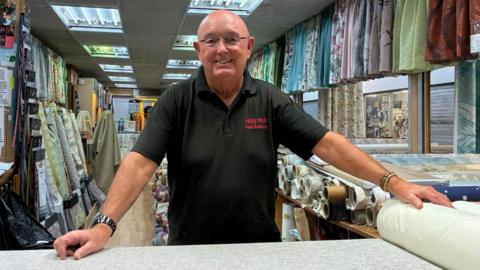 A man wearing glasses and a black polo shirt with Billy Hill Curtains written on is smiling while standing at his stall. He is surrounded by curtain samples.
