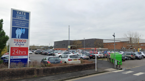 An image of a Yate Shopping Centre sign in front of a car park full of parked cars.