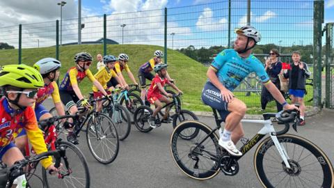 Mark Cavendish wearing blue looking back over his should at children riding the bicycles behind him wearing yellow red and blue.