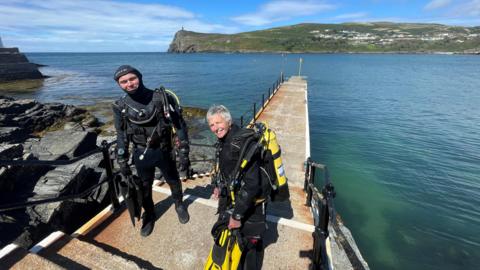 Two people in scuba diving equipment on a jetty in Port Erin on a sunny day.