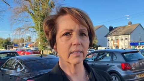 Donna Deeney has short, brown hair and is staring into the camera. She is wearing a black jacket and has silver hoop earrings and a silver necklace. She is standing in front of a number of parked cars.