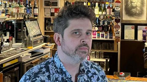 A head and shoulders image of Tom Lagden. He is wearing a black and white floral shirt and sitting in front of a pub bar. 