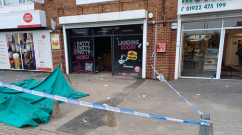 Police tape is in place outside the takeaway which is in between two other businesses. The takeaway has black windows on either  side of its entrance with burger adverts visible. There is a wide concrete pavement outside the shop with green tarpaulin on the ground by the police tape that sections off access to the shop form the pavement.
