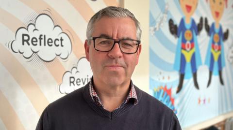 A man in a black jumper standing in a school corridor in front of a wall covered with superhero murals. He is wearing glasses and looking at the camera with a straight face