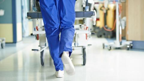 A nurse wearing blue scrubs and white trainers pushes a gurney through a hospital corridor