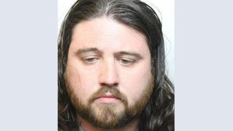 A custody image of Lexi Secker, formerly Alexander Secker. They have long hair and a dark beard