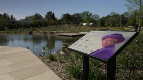 A commemorative information plaque titled "A Garden for a Queen" stands in the foreground, featuring a portrait of Queen Elizabeth II. Behind the sign is a garden landscape including a still pond, stone walkways, and a variety of trees and shrubs.