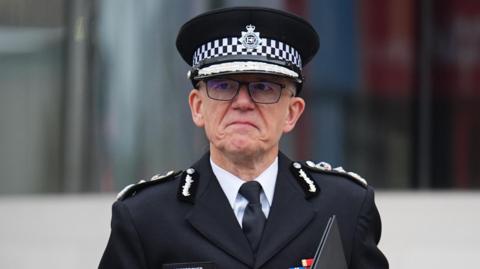 Sir Mark Rowley, Metropolitan Police Commissioner, in uniform with a peaked cap, holding a black folder and has a a serious expression.