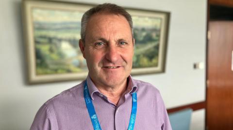 A man with short black-grey hair and a red and white chequered shirt wearing a blue lanyard in front of a blurred wall with a picture hanging on it.