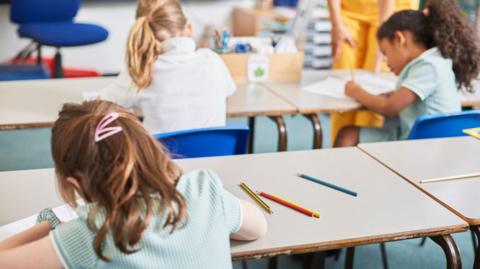Schoolchildren sit at desks in a classroom, writing on paper with pencils. A teacher stands nearby, while coloured pencils lie on the tables in the foreground.