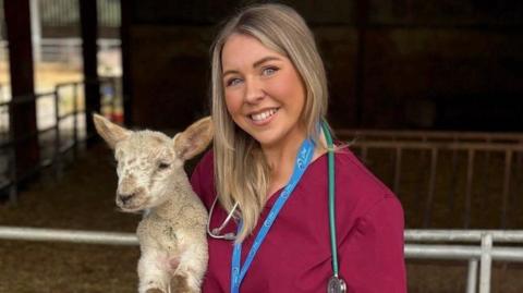 Karis Ramsay holding a lamb in front on an animal pen. Karis is wearing burgundy scrubs, a blue lanyard and ID as well and a stethoscope
