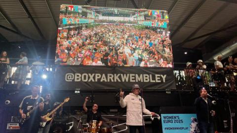A man wearing a white jacket and a hat, talks into a mic on a stage, surrounded by several band members of Supernova - behind them is a big screen showing a large crowd.