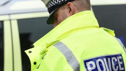 A man wearing a police jacket with police written on the back. He is wearing a hat and standing by a police car