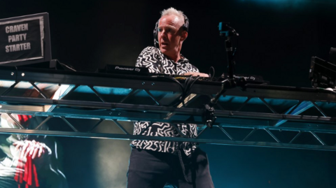 The image shows a DJ performing on stage at a live music event. The DJ is standing behind a large metal-framed table with professional DJ equipment, including mixers and turntables. There is a laptop or controller on the left side with a visible sticker or label that reads “CRAVEN PARTY STARTER.” The DJ is wearing a patterned black-and-white top and dark trousers. Overhead stage lighting creates blue and white spotlights shining down and some haze or smoke effects in the background.