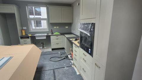 An olive green kitchen with black worktops and large gold handles which is half-renovated and built with protective polystyrene and cardboard around some parts of it. There is a window at the back above the sink.