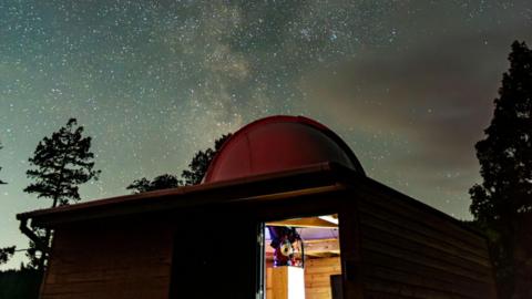 Part of Grizedale Observatory. The building has a dome on its roof with shimmering stars seen in the night sky above - the Milky Way.