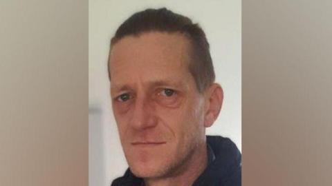 Head and shoulders shot of Vadims Zmicerevskis who has short hair and is wearing a black zipped up jacket and coat. There is a beige wall in the background.