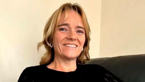 Sandie Peggie, a woman with shoulder-length blonde hair, smiles at the camera while sitting on a couch. She is wearing a black top.