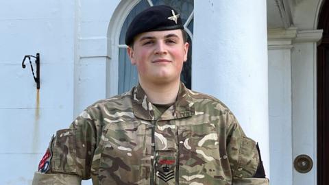 A young person in an army uniform stood in front of a whitewashed building.