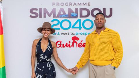 Meagan Good (R) in a black-and-white jumpsuit and brown fedora, and Jonathan Majors (L) in a yellow jumper and beige slacks hold hands as they pose for the camera in front of a sign that reads: "Programme Simandou 2040." A Guinea flag hands furled on the far right of the frame