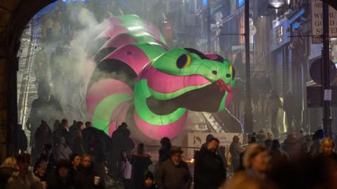 Shows a large illuminated snake in green, pink and black going down Shipquay Street, with people in the foreground in front of an arch.