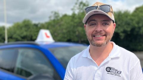 A man with a short grey beard stood in front of a blue car with a learner driver sign on the top of it. He is wearing a beige cap with sunglasses on his head and a white polo top which has the words 'able automatic driving school' stitched on it. He is smiling and looking right at the camera. 