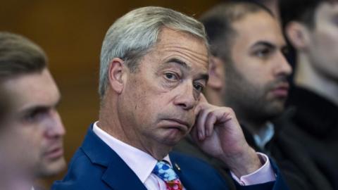 Nigel Farage wears a bright blue suit, pink shirt and blue patterned tie, looking off camera. He is puffing his cheeks.