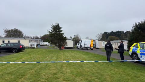 Police officers standing on grass at a caravan park. Police tape cordons off the scene. Police vehicles are also visible. 
