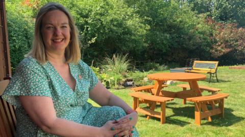 A woman with shoulder length blond hair is wearing a green and white flowered dress. She is sitting by a shed in a sunny garden smiling at the camera