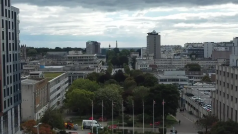 Urban landscape featuring high-rise and smaller buildings, with a green park and trees in the foreground. Several flagpoles with flags are present. The sky is overcast, and vehicles and people are visible throughout the scene.