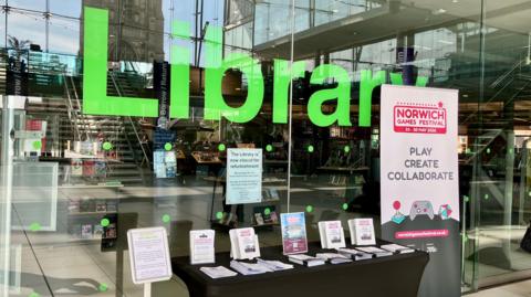 A view of the outside of the library in Norwich. It has a glass window and a sign on it telling people the library is closed. Books and staircases can be seen inside and there is a table outside with a range of leaflets on it.