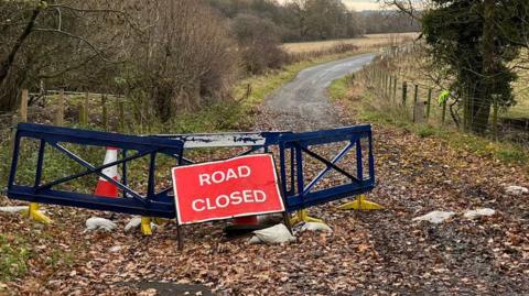 Plastic road closed barriers partially block the entrance to a leaf-strewn road. One of the barriers has been moved to the side of the road.