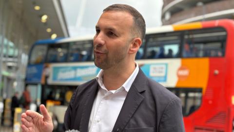 Zack Polanski speaking to someone to the left of the camera. He is mid-speech and is gesticulating with his hand. He has dark, short hair and is wearing a white shirt and grey blazer. Behind him there is a blue, white, orange and red double decker bus which is out of focus.